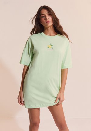 Donna con capelli castano lunghi che indossa un abito maglietta verde chiaro morbido con una piccola grafica di limone, in piedi davanti a uno sfondo beige uniforme.