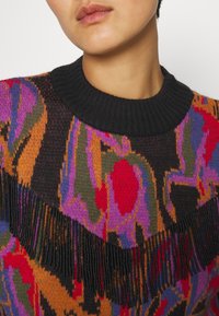 Pull en tricot arborant un motif vibrant en rouge, rose, vert et orange, avec un col noir et un détail de franges perlées le long de la poitrine.