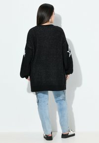 Pull en maille noir surdimensionné avec des motifs d'étoiles sur les manches, manches longues et ourlet côtelé, associé à un jean bleu clair et des chaussures noires.