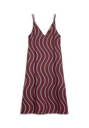 Mujer con largo cabello rojo, vestida con un vestido burdeos sin mangas con rayas onduladas azules y botas negras, de pie frente a un fondo liso.