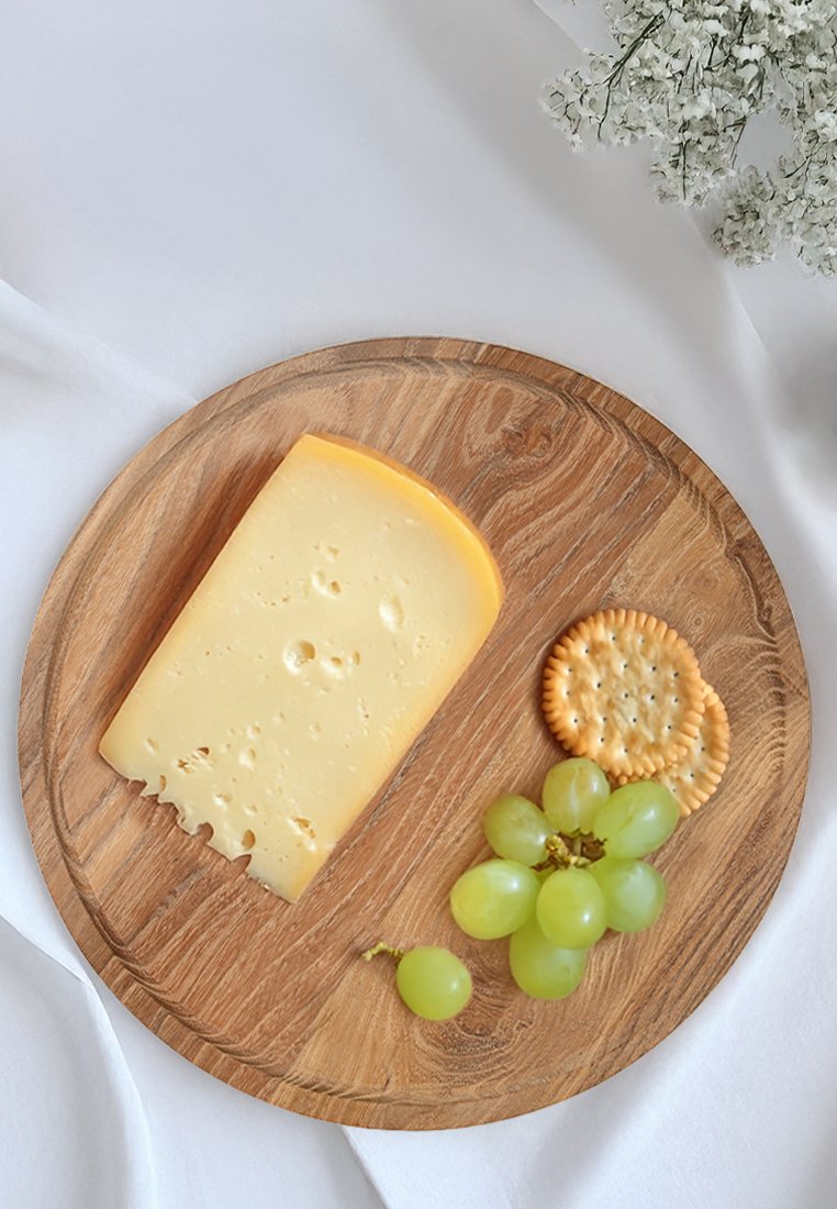 Una rebanada de queso amarillo con agujeros sobre una bandeja redonda de madera, acompañada de uvas verdes y galletas redondas. El fondo es blanco.