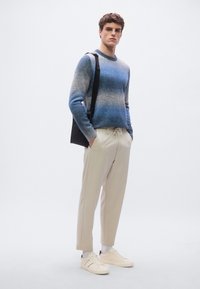 Sininen liukuvärinen neule pyöreällä kaula-aukolla, beige kapenevat housut ja valkoiset lenkkarit. Malli seisoo yksiväristä taustaa vasten kädet taskuissa.