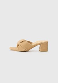 Mule-uri din suede bej cu toc pătrat, cu un design cu curea încrucișată, vârful deschis și o suprafață texturată. Tocul are o înălțime de aproximativ 3 inci.