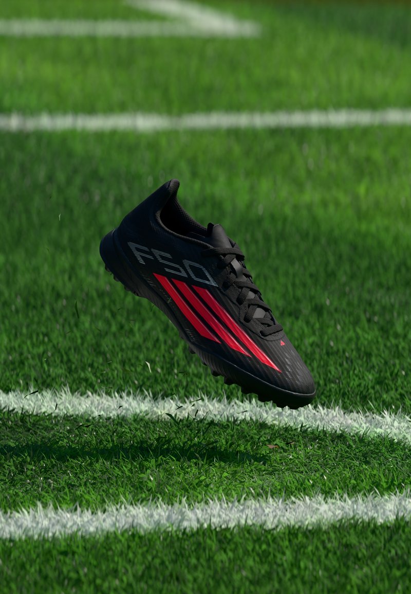 Chaussure de football noire et rouge avec l'inscription "F50" sur un terrain de gazon vert près des lignes blanches de délimitation.