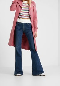 Abrigo de trench de cuero rosa con cintura ceñida, combinado con un suéter de punto a rayas y jeans acampanados de color azul oscuro, acentuado con botas blancas hasta el tobillo.