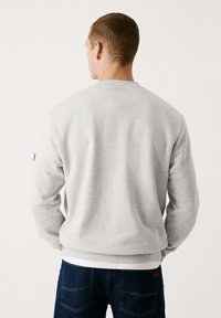 Sweatshirt gris à col rond, avec des poignets et un ourlet côtelés. Présente une accent bleu sur la manche gauche. Porté avec un jean noir.