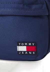 Borsa di tessuto blu con un'etichetta logo rettangolare recante il testo "TOMMY JEANS" e blocchi di colore rosso, bianco e blu. Dettaglio della superficie testurizzata.