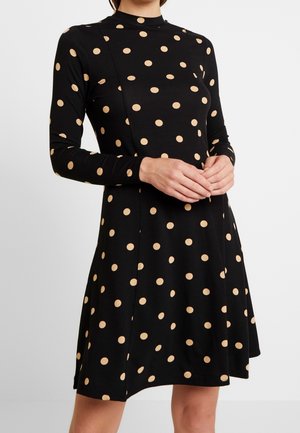 Kvinde iført en sort langærmet kjole med beige polkaprikker, stående med hænderne foldet foran mod en ensfarvet baggrund.