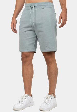 Man draagt lichtgrijze shorts met trekkoord en witte sneakers, staand tegen een effen achtergrond.