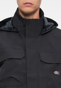 Black jacket featuring a high collar, front zip closure, two large flap pockets, and a logo patch. Textured fabric with a subtle striped pattern.