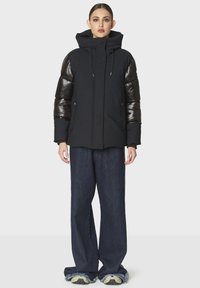 Giacca a vento nera con maniche imbottite lucide e colletto alto. Presenta una cerniera frontale e due tasche laterali. Indossata con pantaloni in denim a gamba larga.