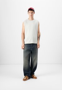 Lichtgrijze mouwloze top, losse donkerblauwe jeans en bruine sneakers met een licht patroon. Model draagt een rode pet. Eenvoudige achtergrond.