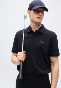 Μπλε ναυτικό πόλο με γιακά, με διακριτική υφή και λογότυπο "BOSS"· το μοντέλο κρατά ένα ρόπαλο γκολφ, φορά καπέλο σε αντίστοιχη μπλε απόχρωση.