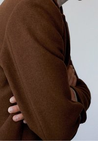Brown wool coat with a structured design, visible seams, and textured fabric, worn with arms crossed in a neutral setting.