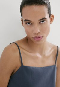 Persona joven con el cabello peinado hacia atrás, vistiendo una camiseta sin mangas de color gris oscuro, mirando directamente a la cámara con una expresión neutral.