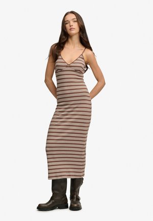 Kobieta o długich brązowych włosach nosi brązowo-białą, w paski, bezrękawnikową sukienkę midi oraz ciemnobrązowe kozaki do kolan, stoi z rękami za plecami.