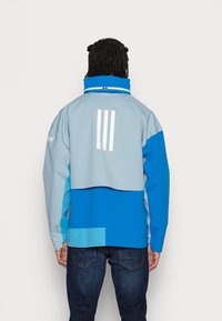 Osoba v modrém a světle šedém saku se třemi vertikálními bílými pruhy na zádech, doplněná tmavě modrými džíny.