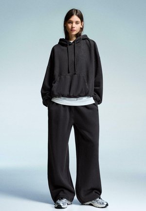 Černá oversized mikina s přední kapsou a šňůrkou, přehozená přes bílou košili. V kombinaci s volnými černými kalhotami a šedými sportovními botami.