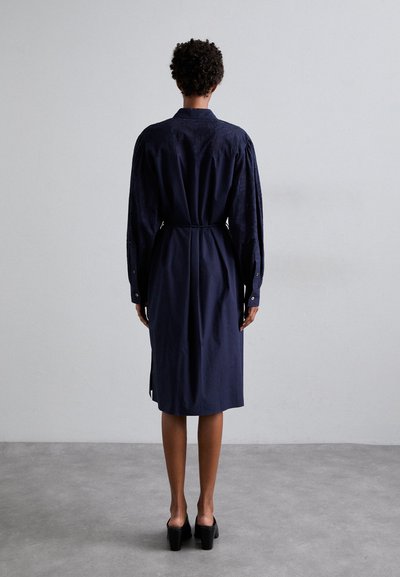 Granatowa sukienka z długim rękawem, z paskiem w talii, o teksturowanym materiale i guzikach przy mankietach. Ma prosty krój i boczne rozcięcia.