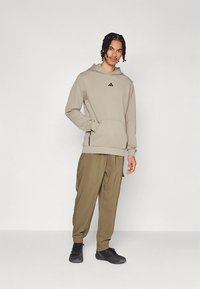 Μπεζ φούτερ με κουκούλα και μπροστινή τσέπη, με μαύρο λογότυπο, συνδυασμένο με φαρδιά cargo παντελόνια σε ελαιώδες πράσινο και μαύρα αθλητικά παπούτσια.