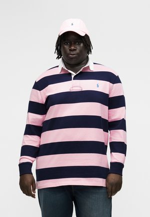 Egy férfi rózsaszín és tengerészkék csíkos rugby pólóban, rózsaszín sapkában és kék farmernadrágban, egyszínű világosszürke háttér előtt áll.