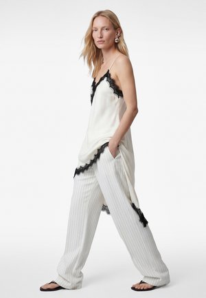 Vrouw loopt, gekleed in witte wijde broek met krijtstrepen, een crèmekleurige slipjurk met kanten rand over de broek, zwarte sandalen en grote zilveren oorbellen.