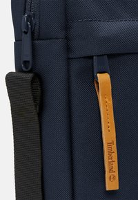 Zaino navy con tessuto testurizzato, una tracolla nera e un tirazip in pelle con la scritta "Timberland" impresso. Dettagli della cerniera in navy.