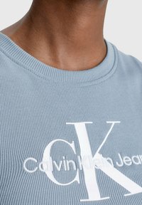 T-shirt en coton ribbed bleu clair avec un grand logo "CK" blanc et le texte "Calvin Klein Jeans" à l'avant, avec un col rond.