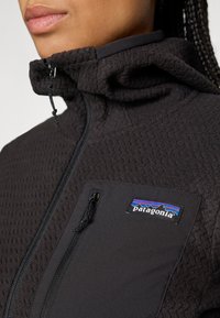 Close-up of person wearing black textured Patagonia jacket with zippered chest pocket and high collar zipped up.