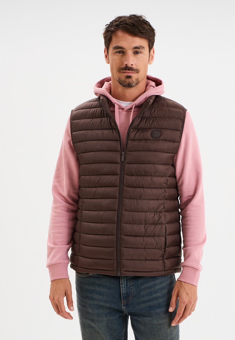 Mann, der einen rosa Hoodie unter einer braunen, gesteppten Weste mit Reißverschluss und blauen Jeans trägt, vor einem schlichten weißen Hintergrund steht.