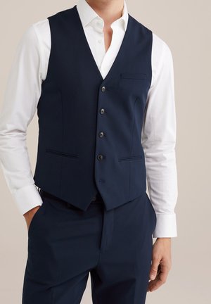 Homme portant un gilet bleu foncé boutonné sur une chemise blanche avec un pantalon bleu foncé, debout avec une main dans la poche.