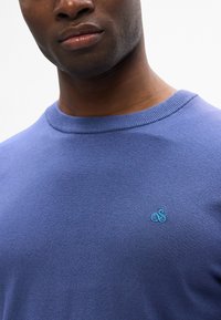 Blauer Baumwollpullover mit rundem Halsausschnitt und gerippten Kanten, versehen mit einem kleinen türkisfarbenen Logo auf der Brust. Glatte Textur und lässiges Design.