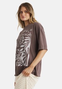 Junge Frau mit blonden Haaren, die ein lockeres braunes T-Shirt mit weißem Blumen- und Mondphasen-Print trägt und einen cremefarbenen Strickrock hält.