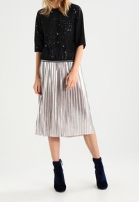 Haut noir à motif moucheté, associé à une jupe plissée argentée métallique et des bottines bleu marine. Texture lisse et silhouette moderne.