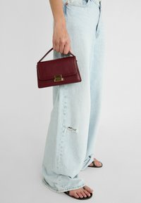Bordeauxfarbenes Lederhandtasche mit einer strukturierten rechteckigen Form, ausgestattet mit einem vergoldeten Verschluss und einem kurzen Henkel, gehalten von einer Person.