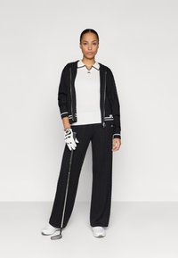 Μαύρο ζακέτα με πλέξη καλωδίων και φερμουάρ, με λευκές ρίγες στους μανίκους, λευκή πόλο μπλούζα με μαύρο γιακά, μαύρο παντελόνι, λευκό γαντί του γκολφ και παπούτσια.