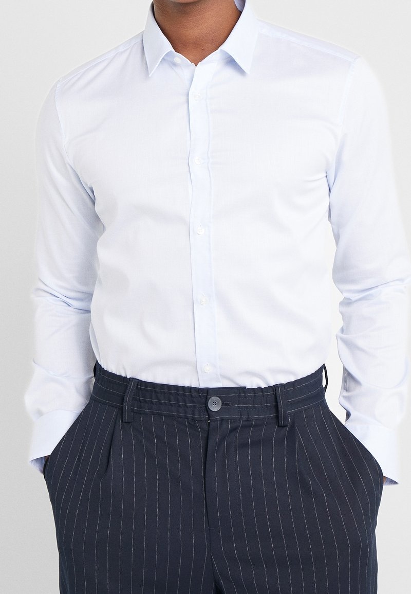 Homme portant une chemise blanche cintrée boutonnée, rentrée dans un pantalon bleu marine à fines rayures, les mains dans les poches.