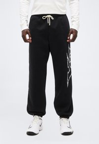 Άνδρας που φοράει μαύρο παντελόνι φόρμας με λευκό κορδόνι και σχέδιο στο πλάι, λευκή μπλούζα με μακριά μανίκια και λευκά αθλητικά παπούτσια με μαύρες λεπτομέρειες.