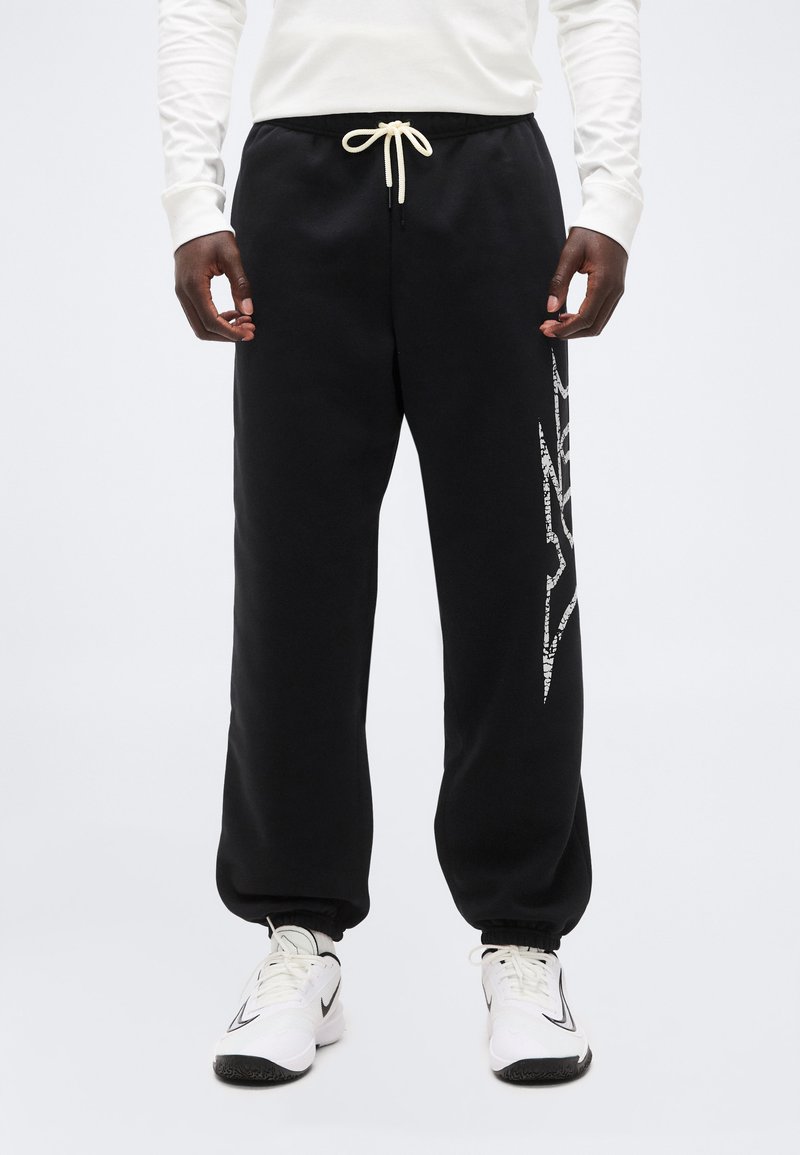 Άνδρας που φοράει μαύρο παντελόνι φόρμας με λευκό κορδόνι και σχέδιο στο πλάι, λευκή μπλούζα με μακριά μανίκια και λευκά αθλητικά παπούτσια με μαύρες λεπτομέρειες.