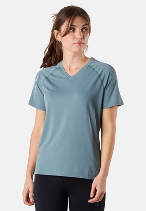 Femme aux cheveux bruns portant un t-shirt bleu clair à col en V et manches courtes ainsi que des leggings noirs, regardant légèrement vers sa gauche.