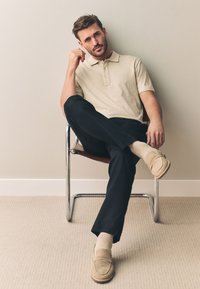 Tricou polo bej, pantaloni negri și mocasini căprui cu detalii de cusătură. Subiect așezat pe un scaun cromat în fața unui perete simplu.