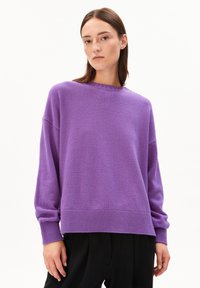 Junge Frau mit glattem braunem Haar, die einen lockeren lila Pullover und schwarze Hosen trägt, vor einem schlichten weißen Hintergrund.