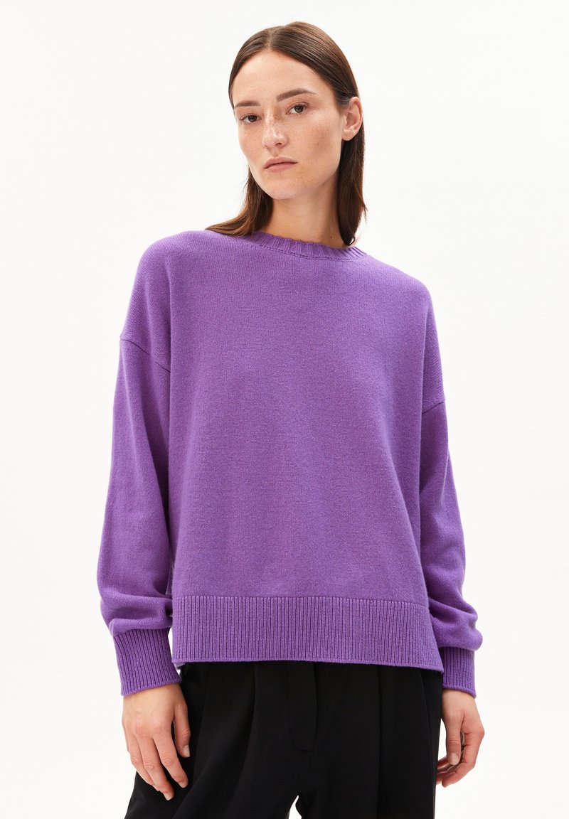 Junge Frau mit glattem braunem Haar, die einen lockeren lila Pullover und schwarze Hosen trägt, vor einem schlichten weißen Hintergrund.