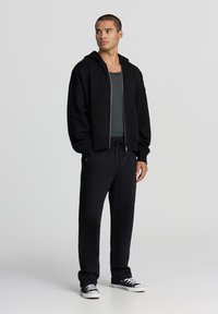 Felpa nera con zip e texture morbida, abbinata a pantaloni da jogging neri con cordino regolabile e sneakers nere.