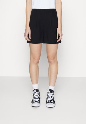 Personne portant un short noir, des chaussettes blanches et des baskets montantes noires, debout devant un fond blanc uni.