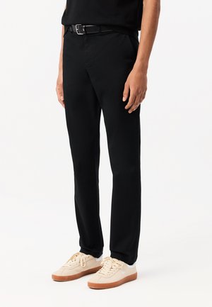 Black trousers with a tailored fit, featuring belt loops and side pockets, paired with light-colored sneakers with a rubber sole.