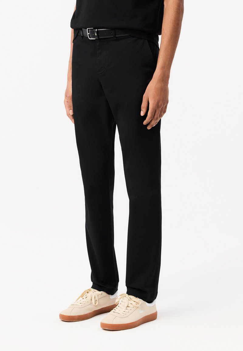 Zwarte broek met een getailleerde pasvorm, voorzien van riemlussen en zijzakken, gecombineerd met lichtgekleurde sneakers met een rubberen zool.