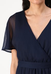 Σκούρο μπλε φόρεμα με V-deck και κοντά, φαρδιά μανίκια. Φτιαγμένο από υφασμάτινη ύλη με υφή, διαθέτει καθορισμένη μέση με ταιριαστή ζώνη.