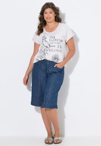 Camiseta gris claro con diseño gráfico negro que presenta olas y un sol. Combinada con culottes de mezclilla oscura y sandalias grises. Un look simple y casual.