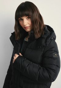 Czarna pikowana kurtka z kapturem, z zapięciem na zamek, o teksturowanym wzorze i z naszywką z logo na lewym rękawie.
